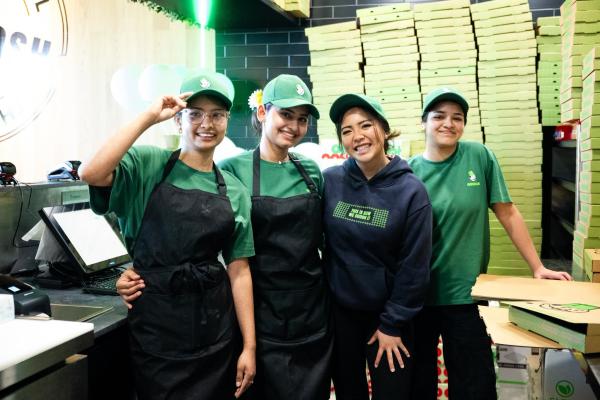 Four smiling OOSHMAN team members in green uniforms inside a busy restaurant kitchen