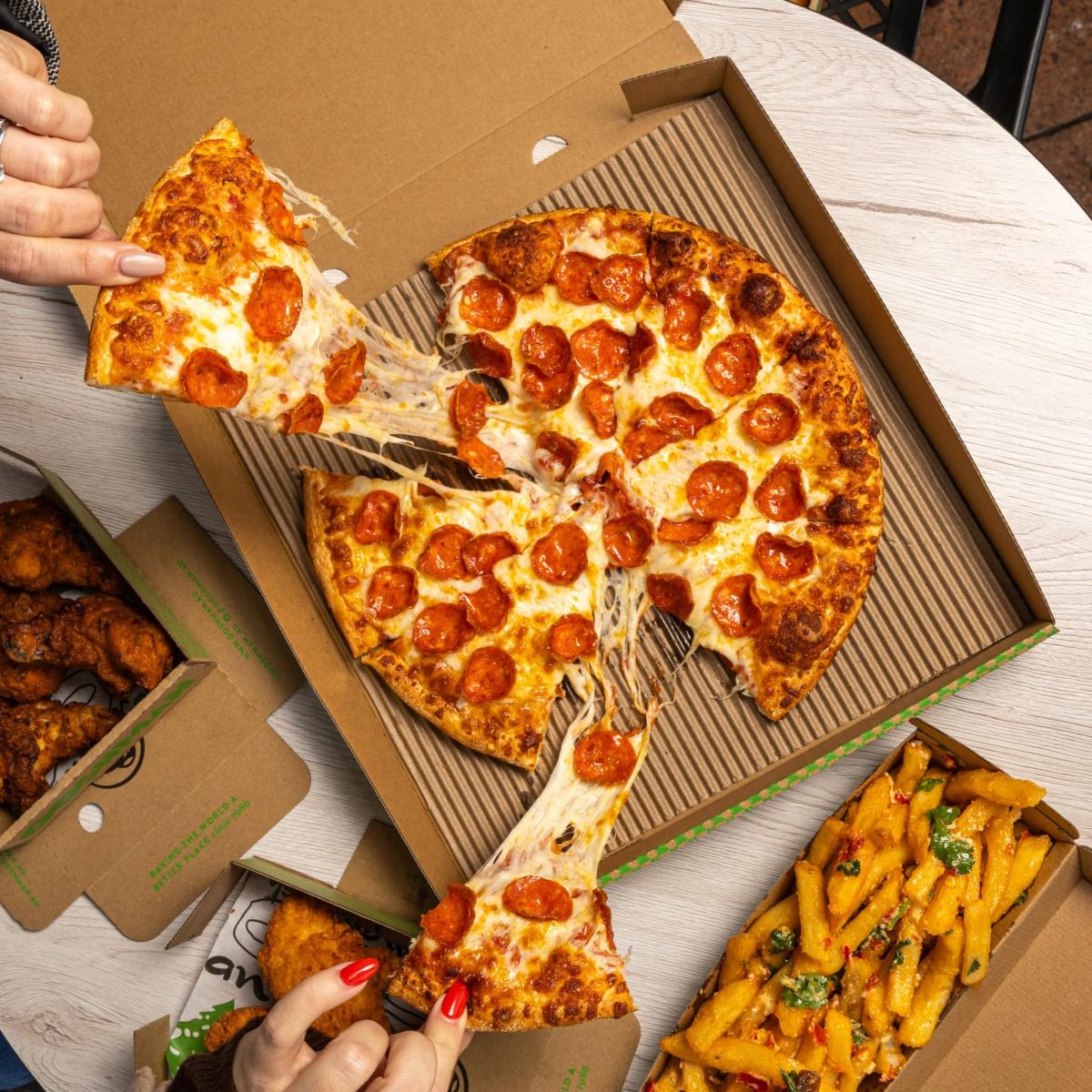 Hands taking slices from a boxed pepperoni pizza with chips and chicken sides on a table