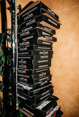 Tall stack of branded pizza takeaway boxes beside a leafy plant and warm textured wall