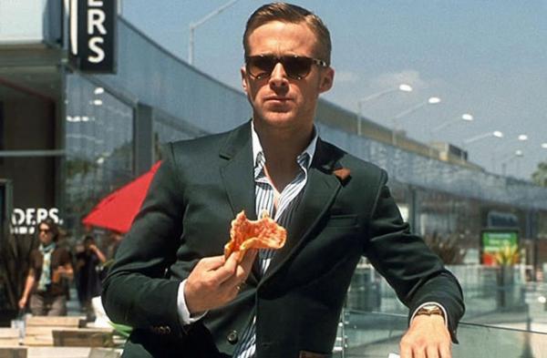 Man in a dark suit and sunglasses holding a slice of pizza in a shopping centre