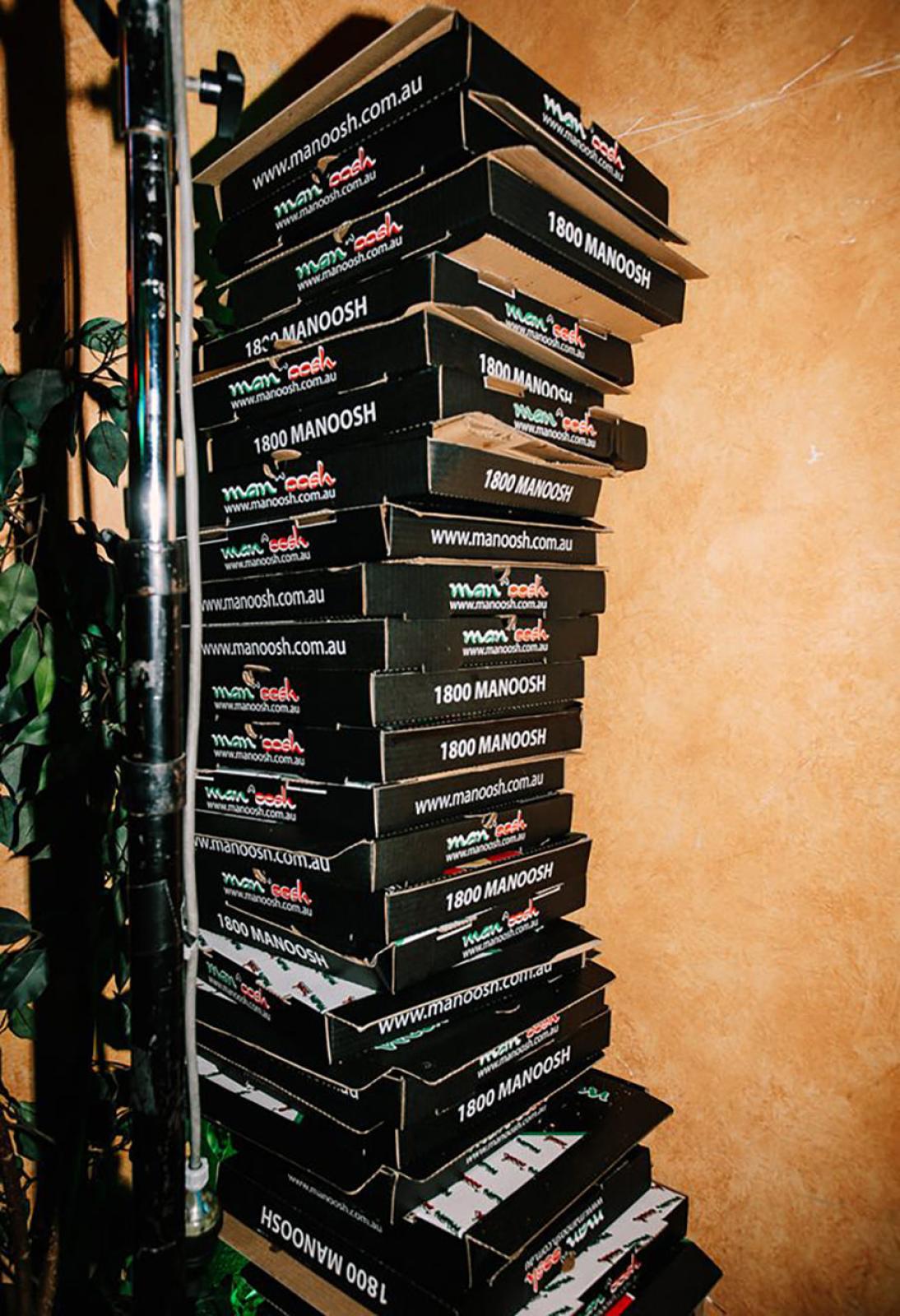 Tall stack of black OOSHMAN pizza boxes beside a leafy plant against a warm textured wall