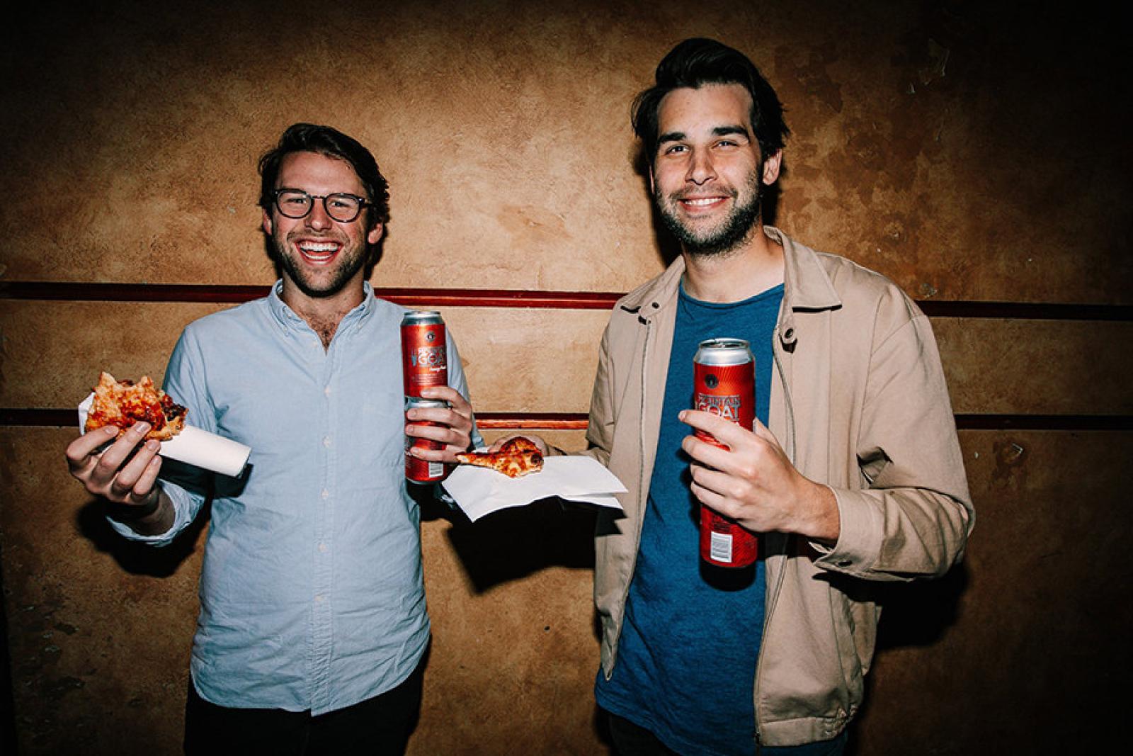 Two men holding pizza slices and canned drinks against a textured brown wall