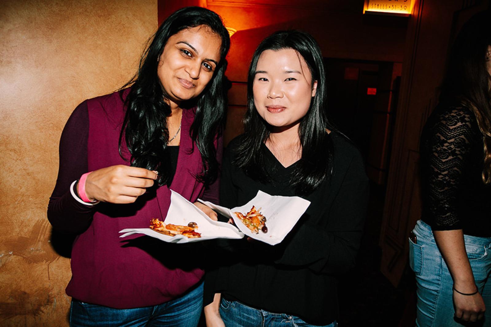 Two women enjoying slices of pizza at an indoor food event