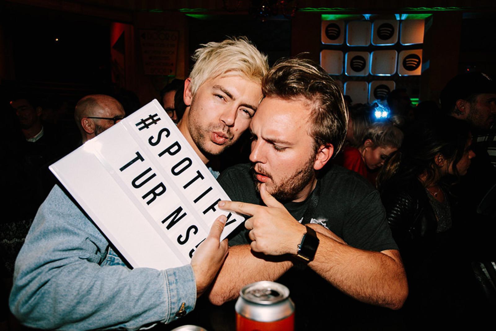 Two men holding a sign reading “#Spotify Turns” at a dimly lit party