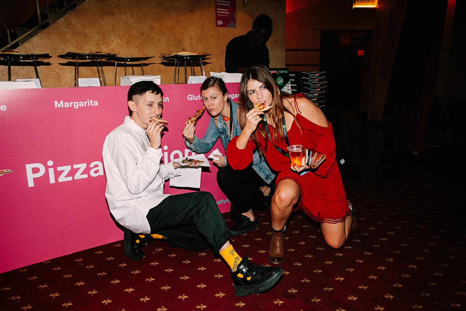 Three friends eating pizza beside a pink pizza counter at a cinema event