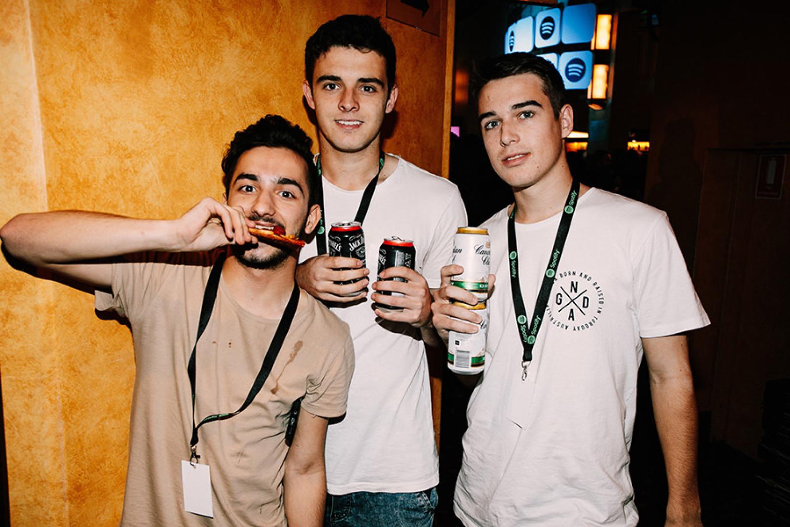 Three young men wearing lanyards hold canned drinks and a snack at an indoor event.