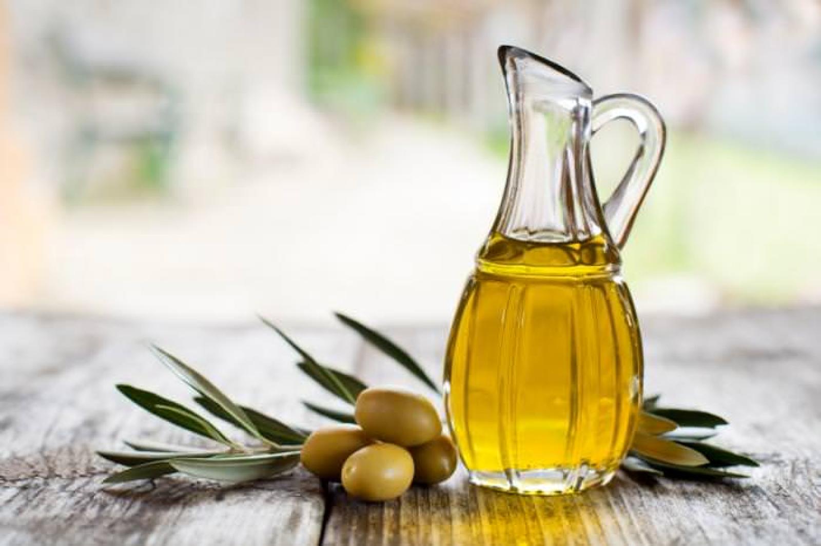 Glass cruet of golden olive oil with green olives and olive leaves on a rustic wooden table