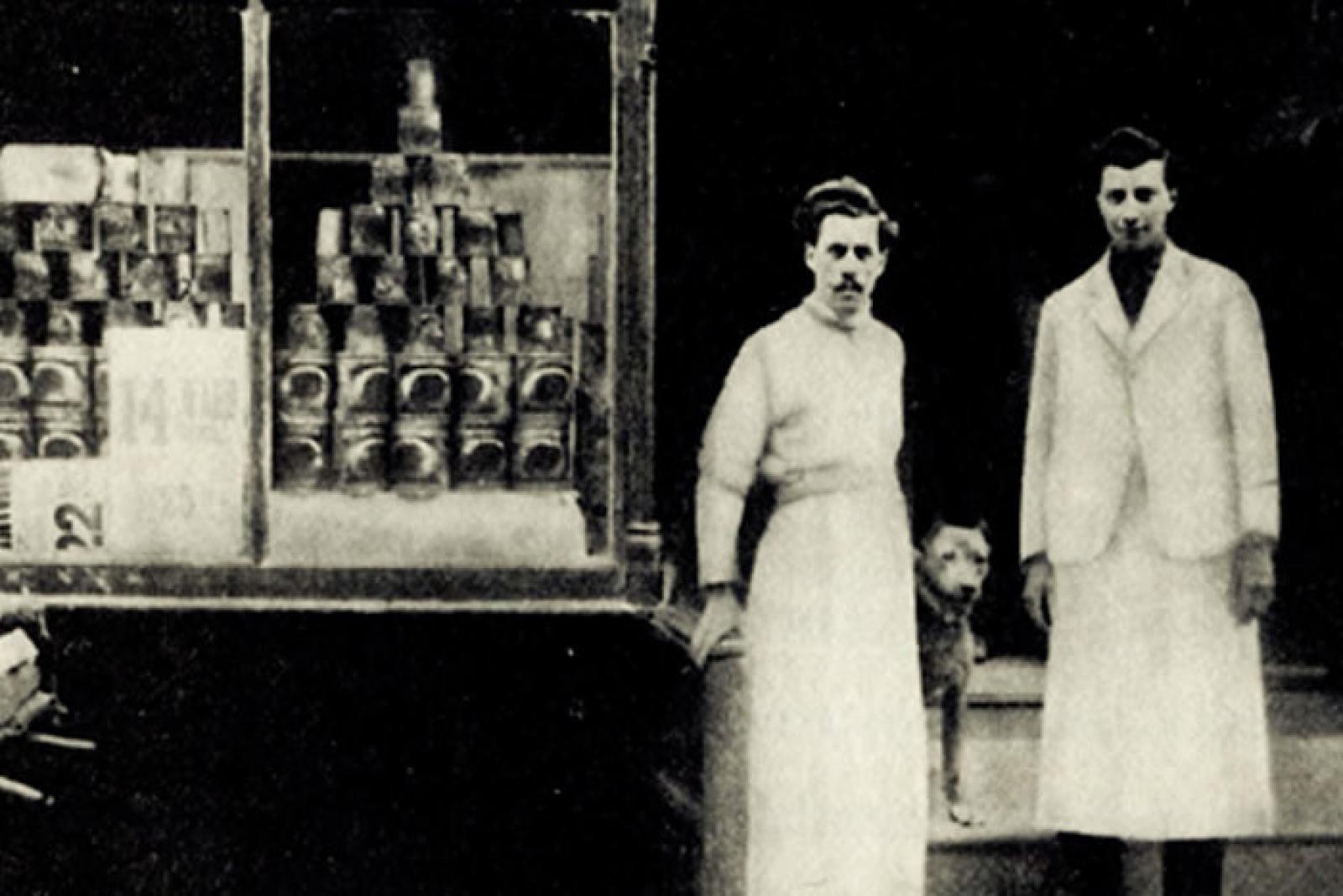 Historic black-and-white photo of two apron-wearing shop workers beside a display window