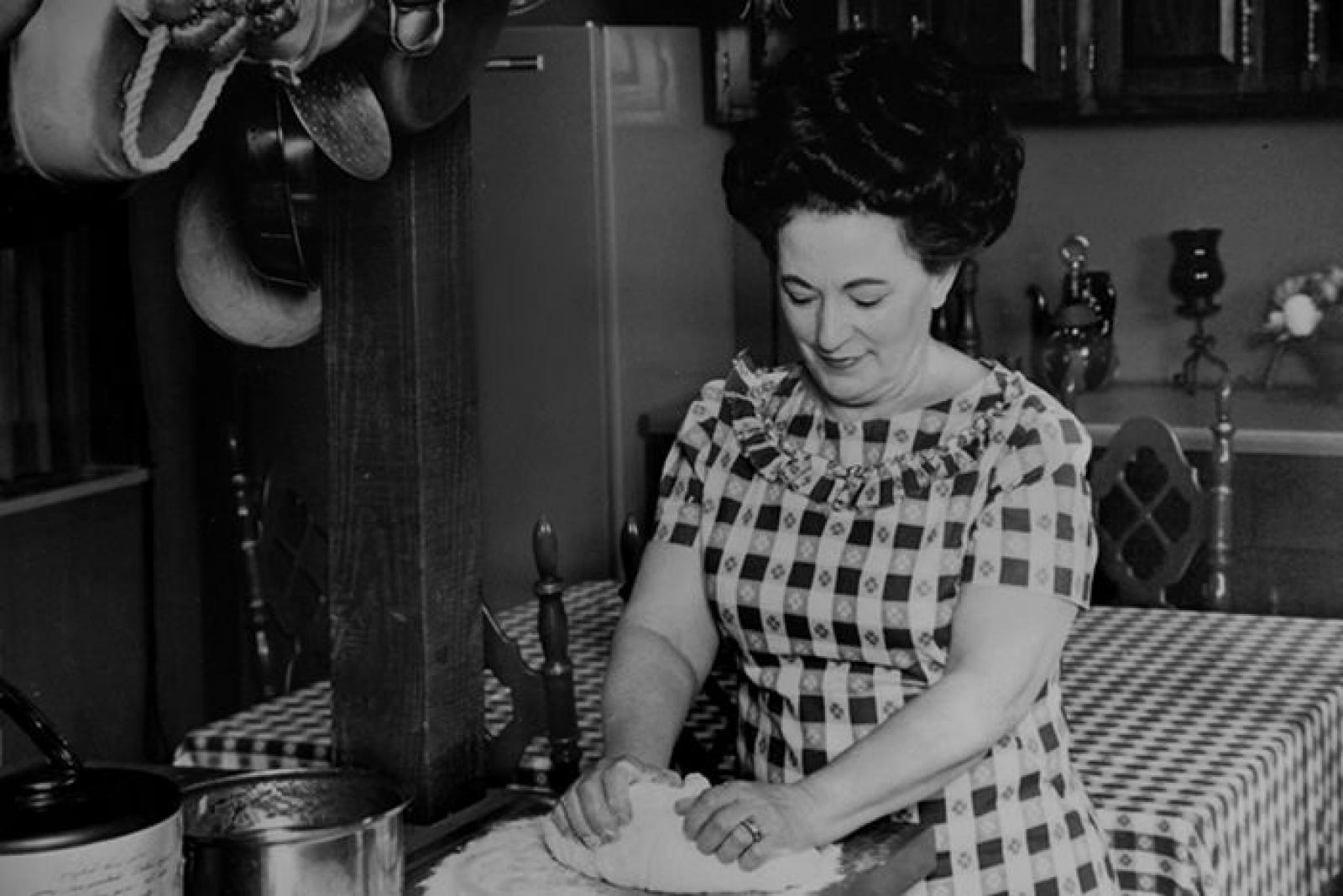 Black-and-white photograph of a woman kneading dough in a traditional home kitchen