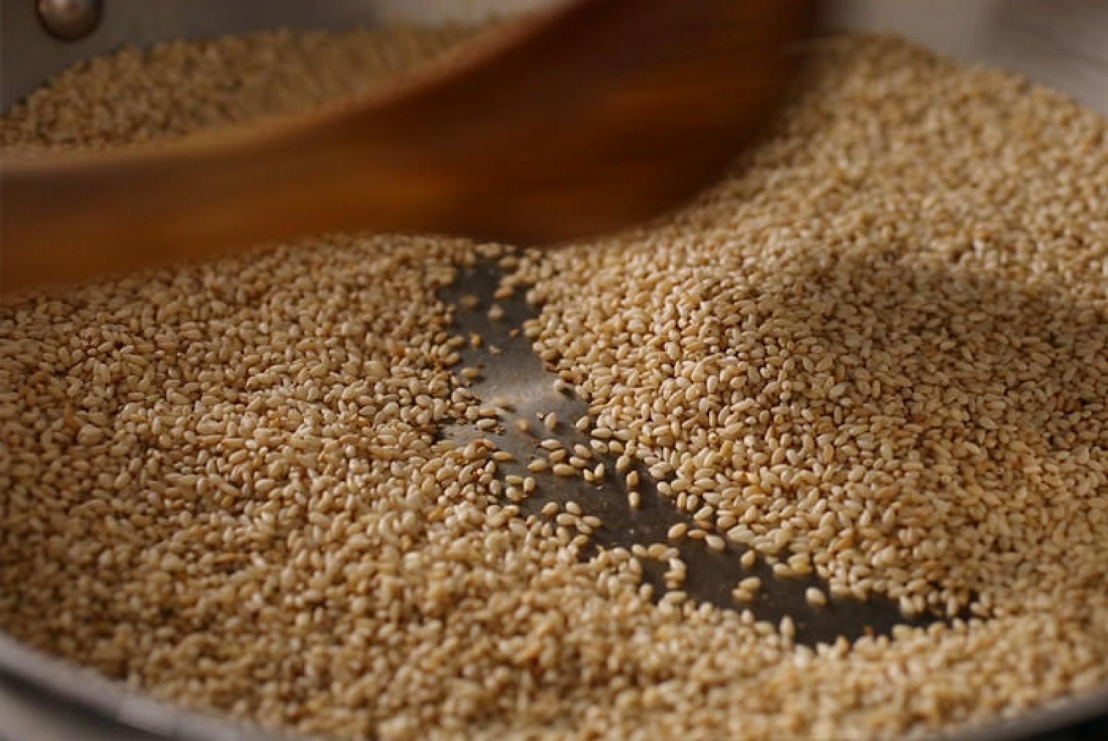 Wooden spoon surrounded by pale sesame seeds on a dark baking tray