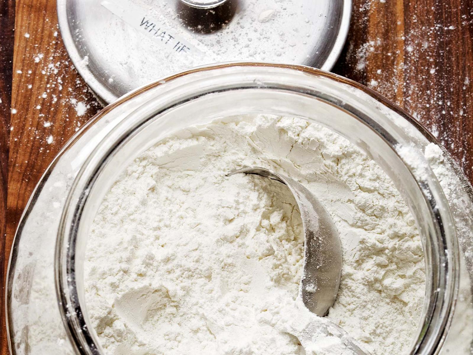 Overhead view of flour in a glass bowl with a metal scoop on a wooden work surface