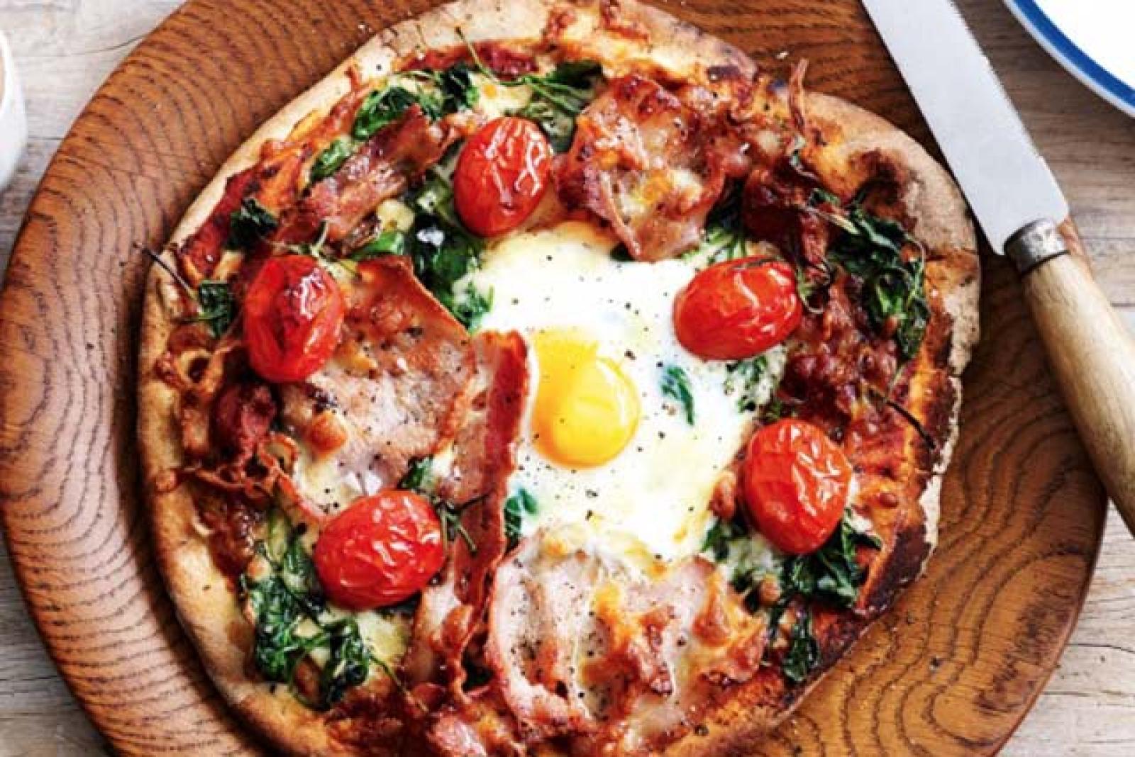 Breakfast pizza with fried egg, cherry tomatoes, spinach and bacon on a wooden board