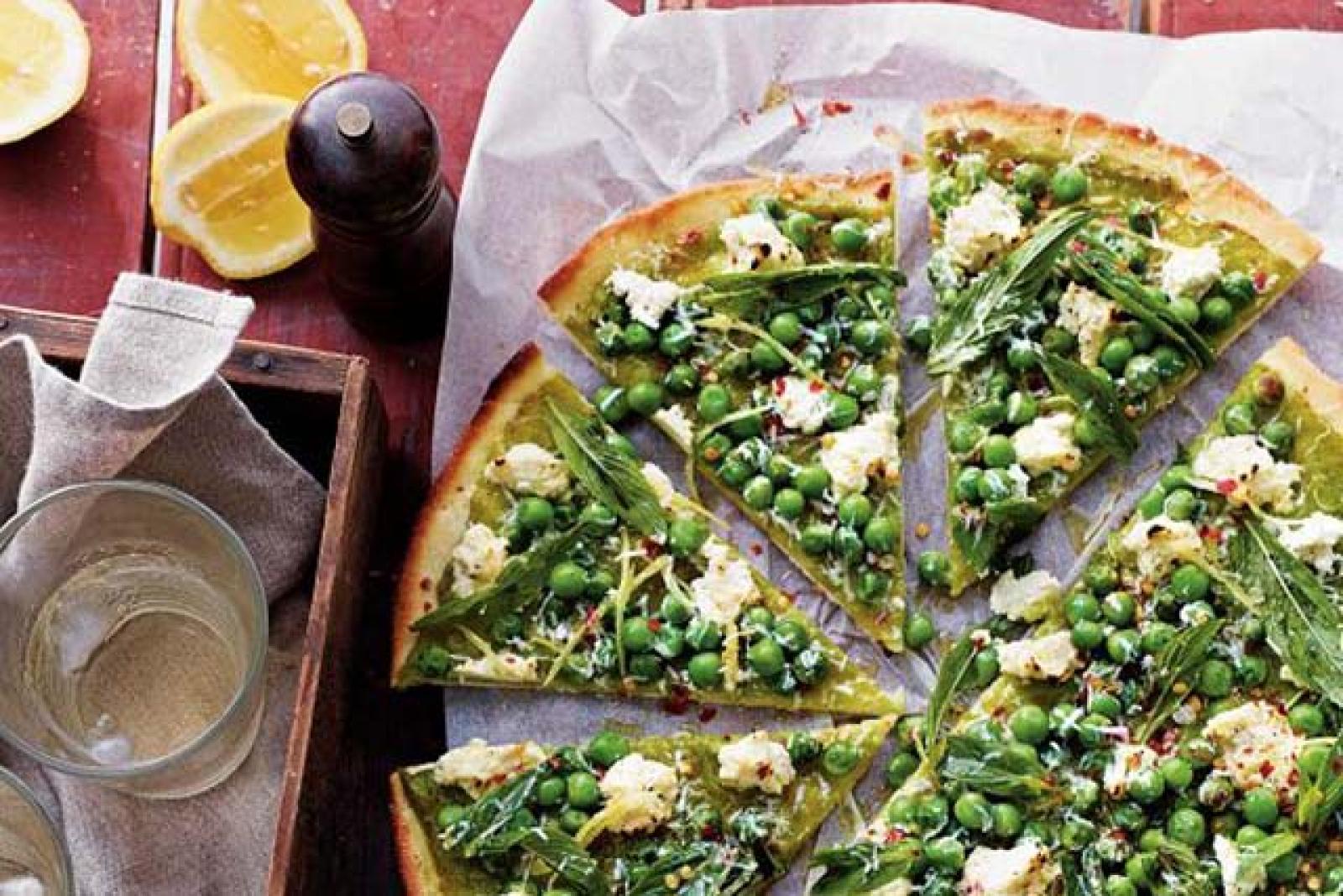 Sliced vegetable pizza topped with greens, peas and crumbled cheese beside lemon wedges