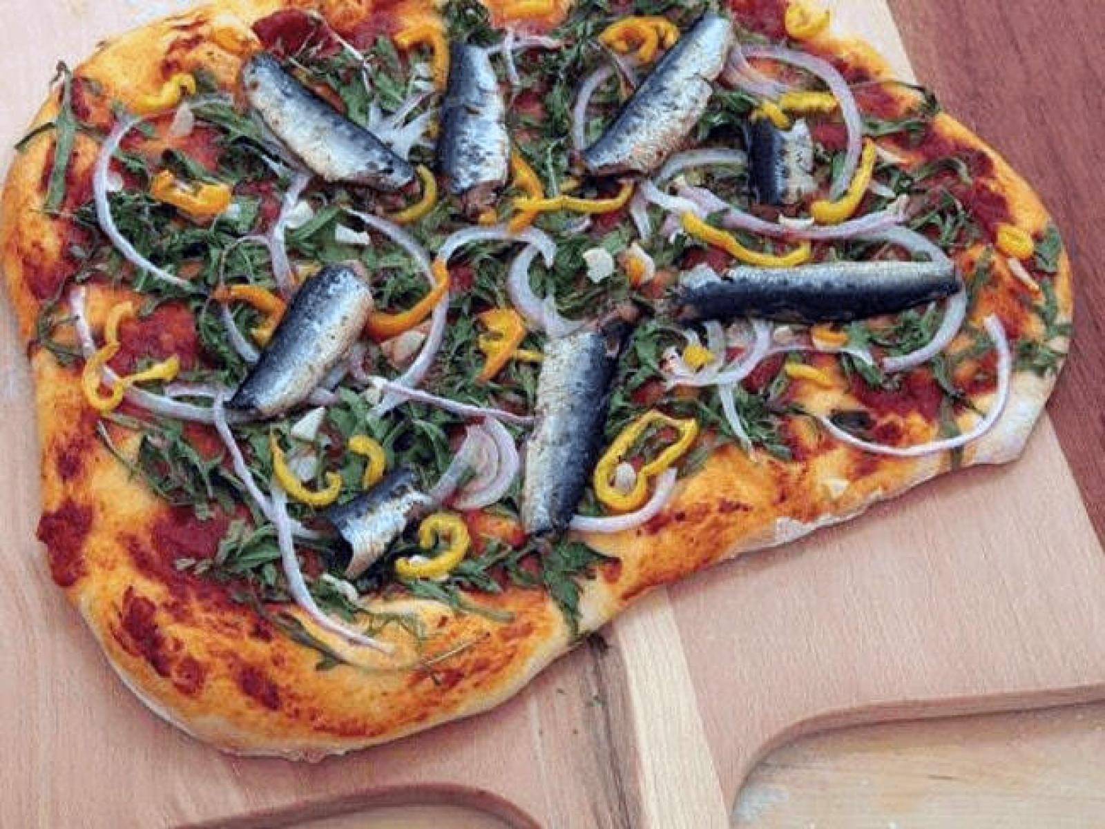 Rectangular pizza topped with whole sardines, olives, onion and peppers on a wooden board