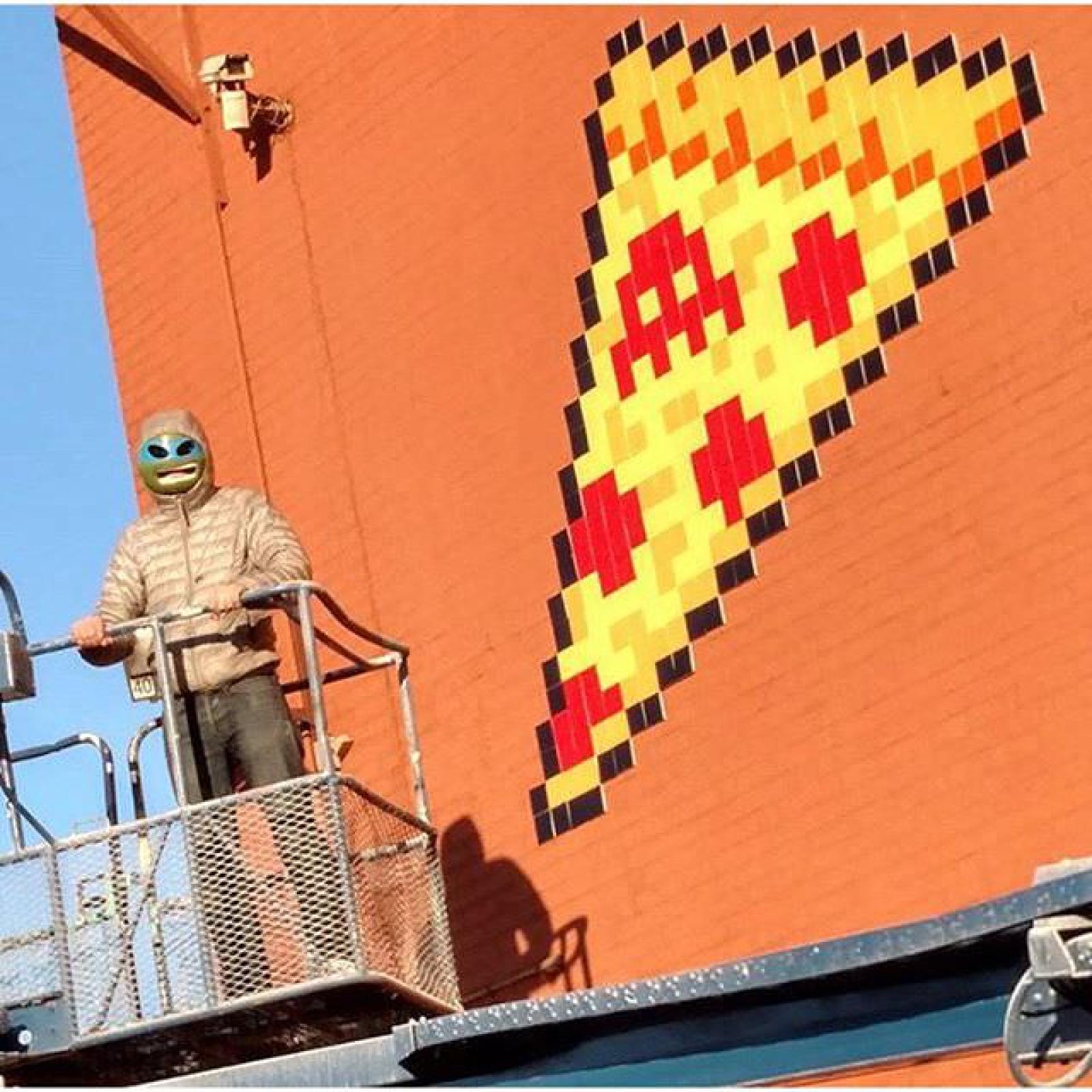 Worker on a lift beside a pixel-style pizza mural on an orange exterior wall