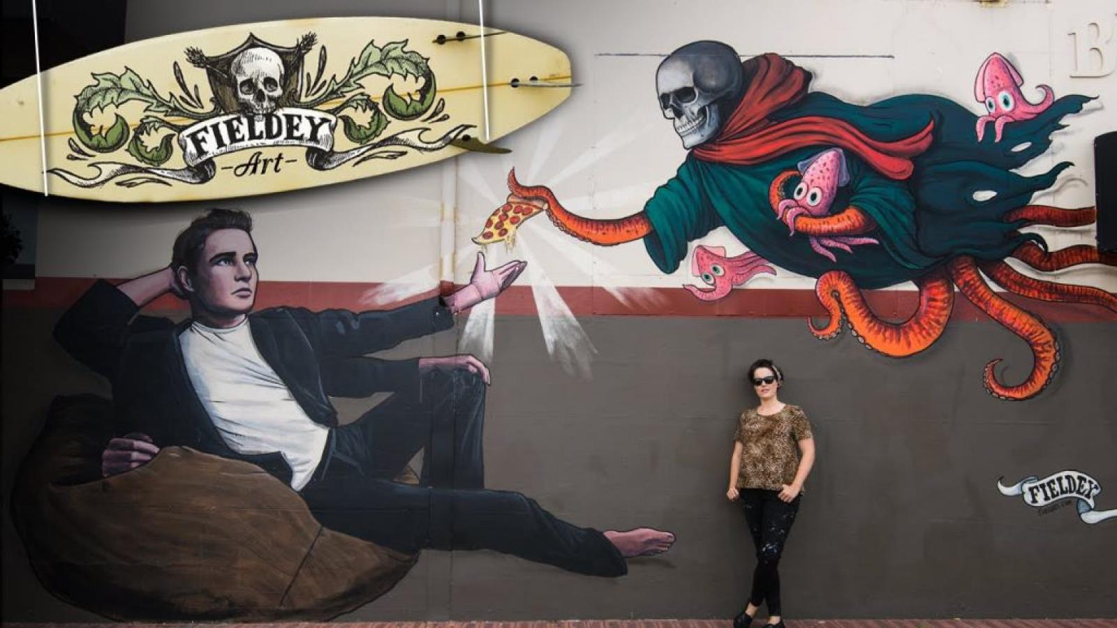 Large illustrated mural of a reclining man and flying superhero figures, with a woman standing below