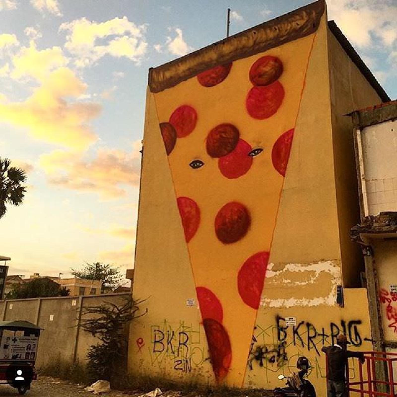 Large pizza slice mural painted on a graffitied building wall at sunset