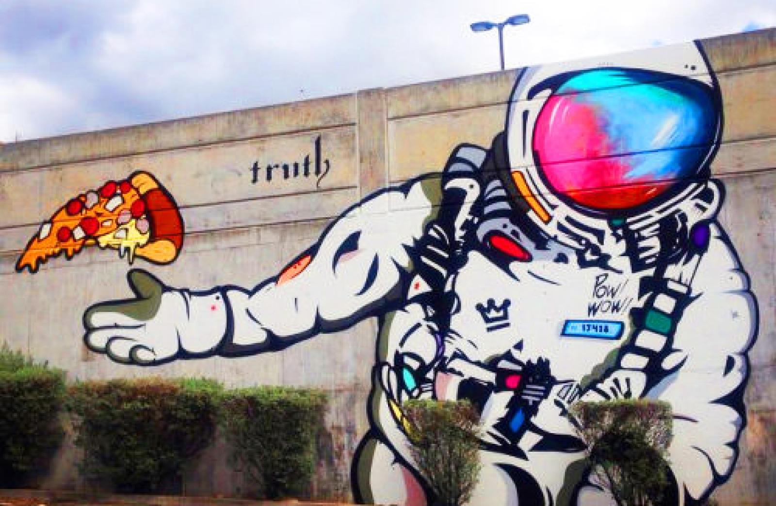 Street mural of an astronaut reaching for a floating slice of pizza