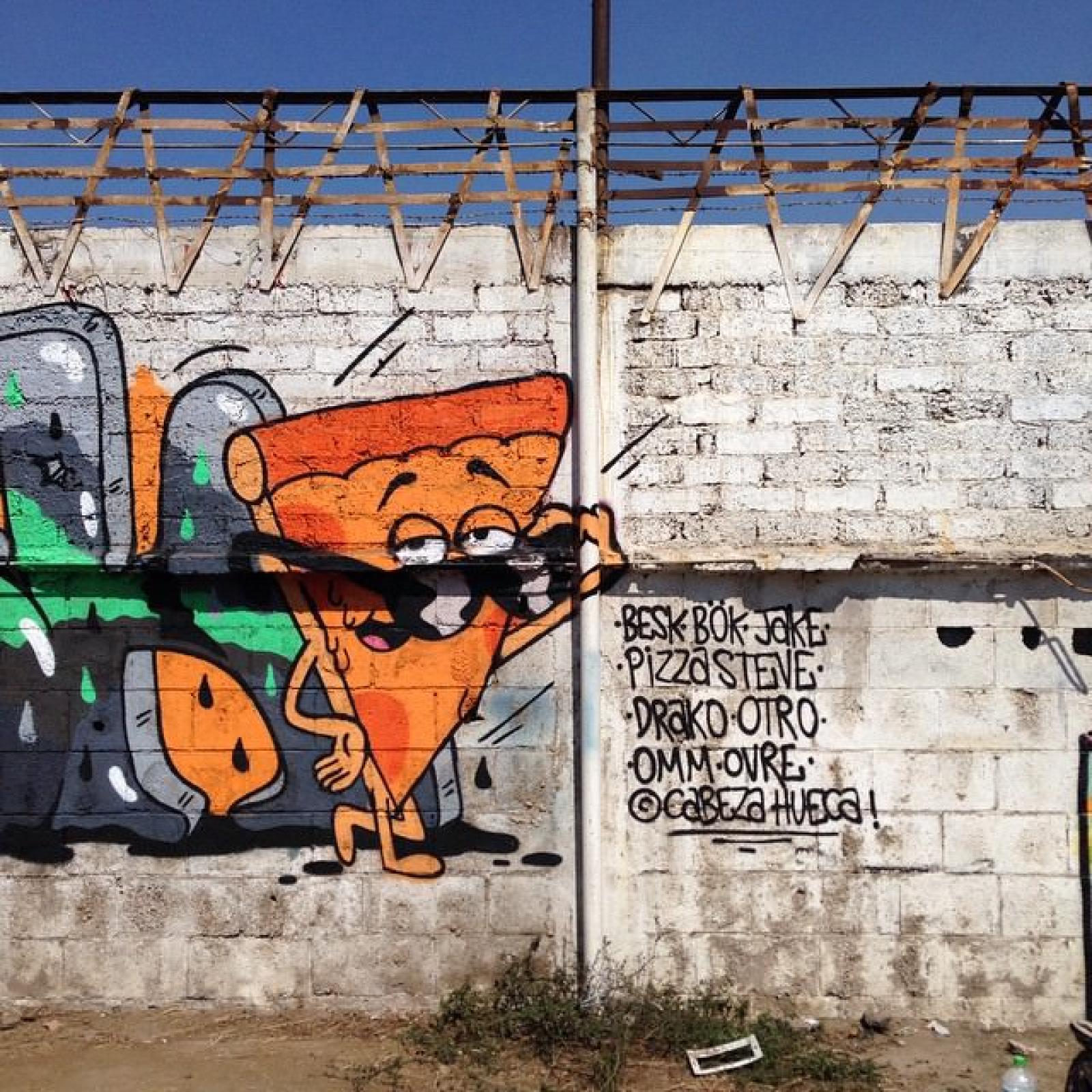 Colourful pizza slice graffiti mural on a white brick wall beneath a rusted metal awning