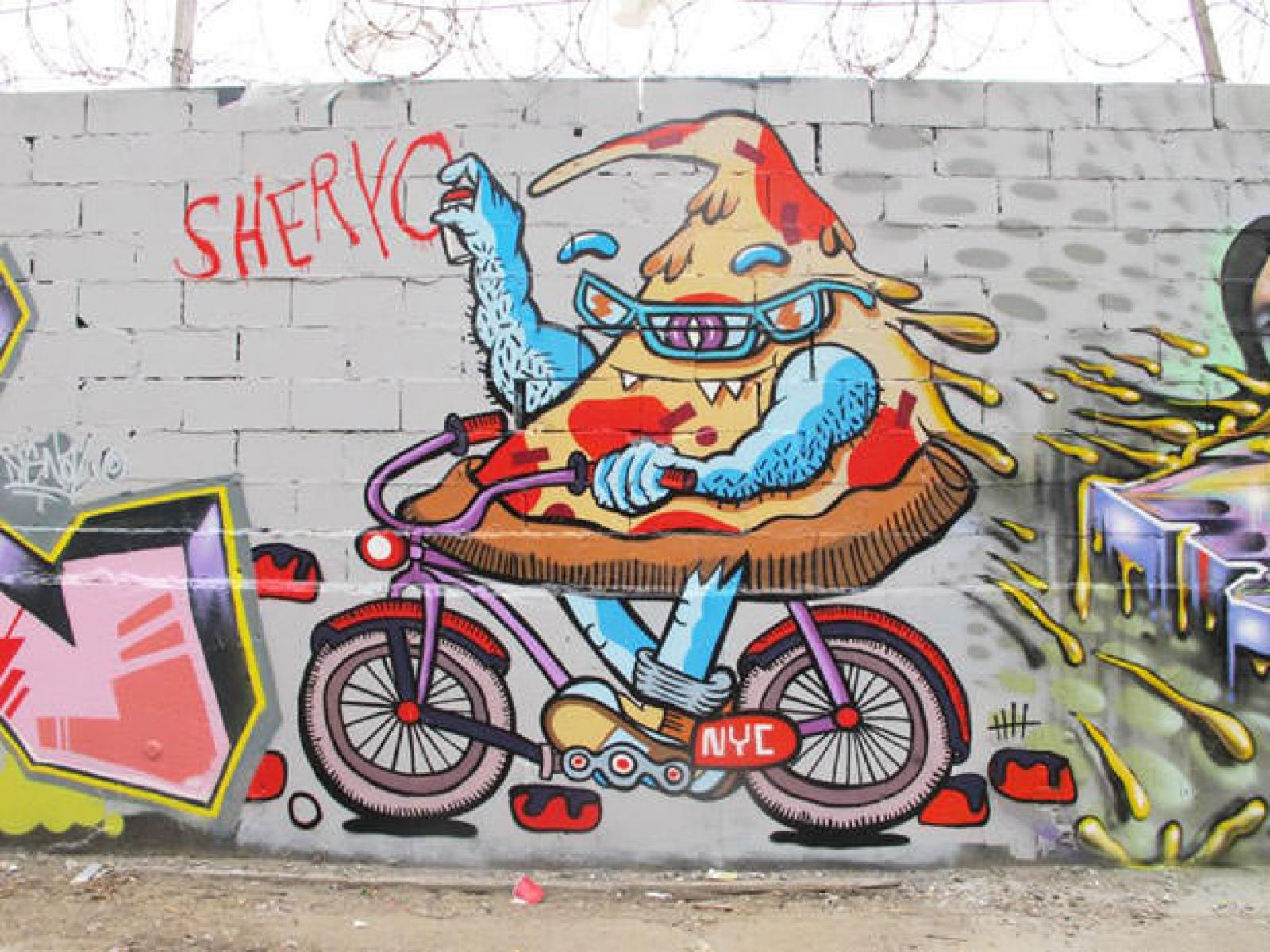 Colourful street-art mural of a cartoon pizza character riding a red bicycle