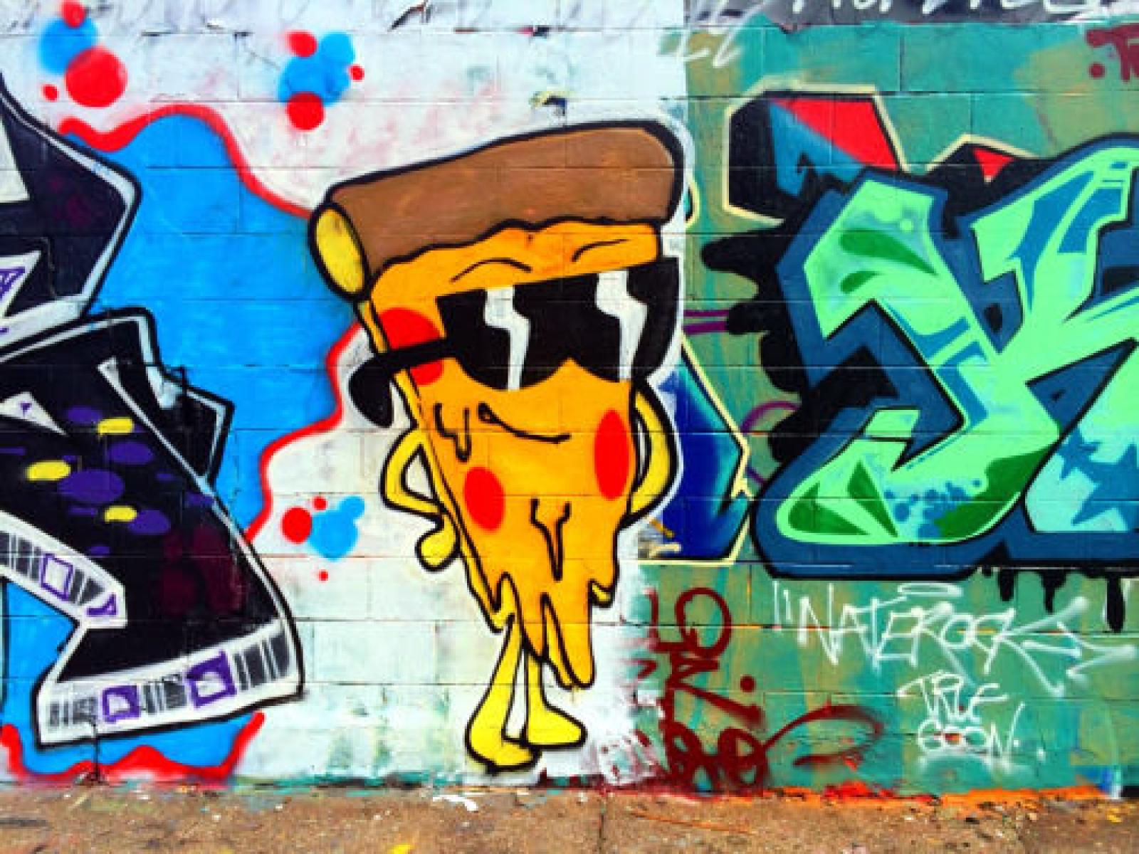 Colourful street mural of a smiling pizza slice wearing sunglasses beside bold graffiti
