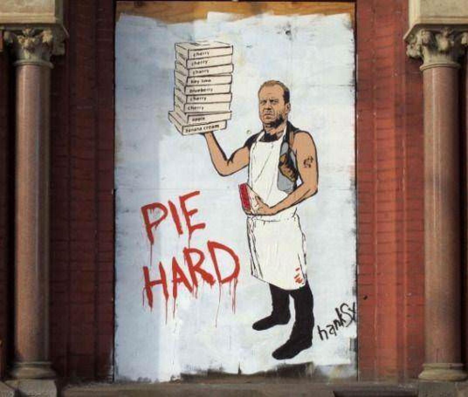 Street-art mural of an apron-wearing man holding pizza boxes beside the words “Pie Hard”.