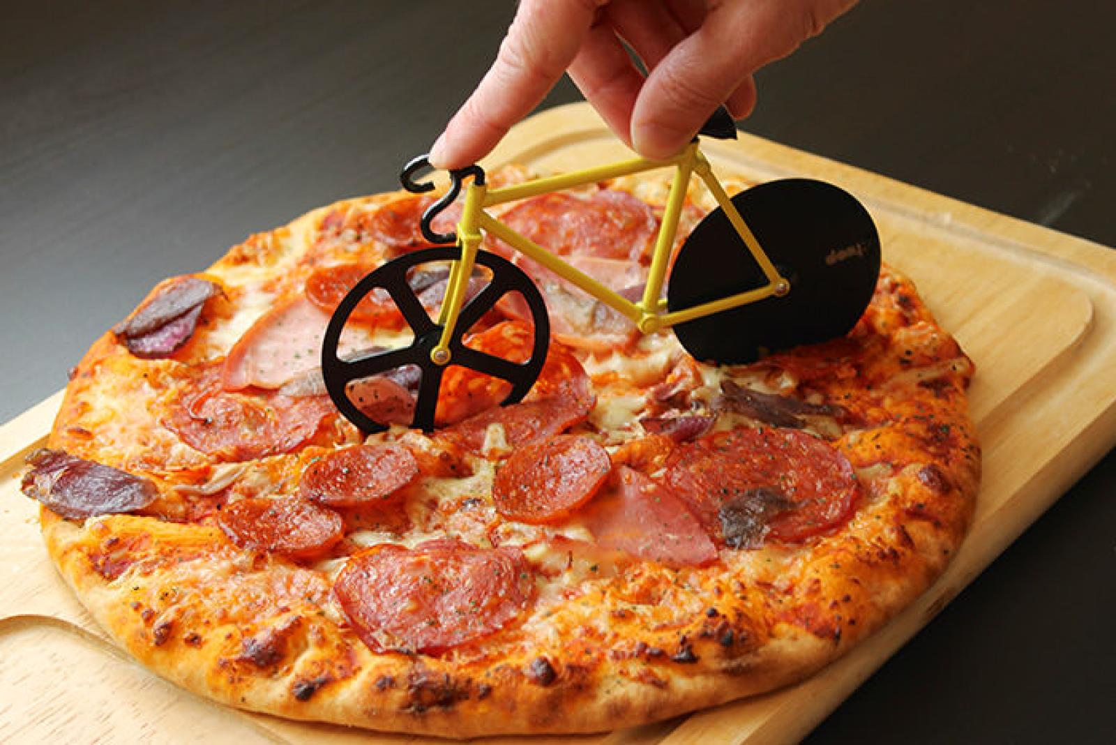 Hand using a bicycle-shaped pizza cutter to slice a pepperoni pizza on a wooden board