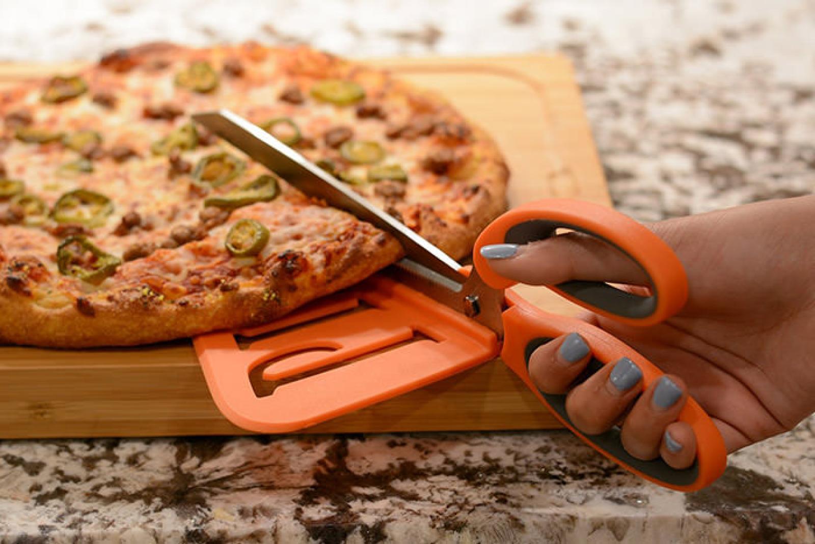 Hand using orange pizza scissors to cut a fresh herb-topped pizza on a wooden board