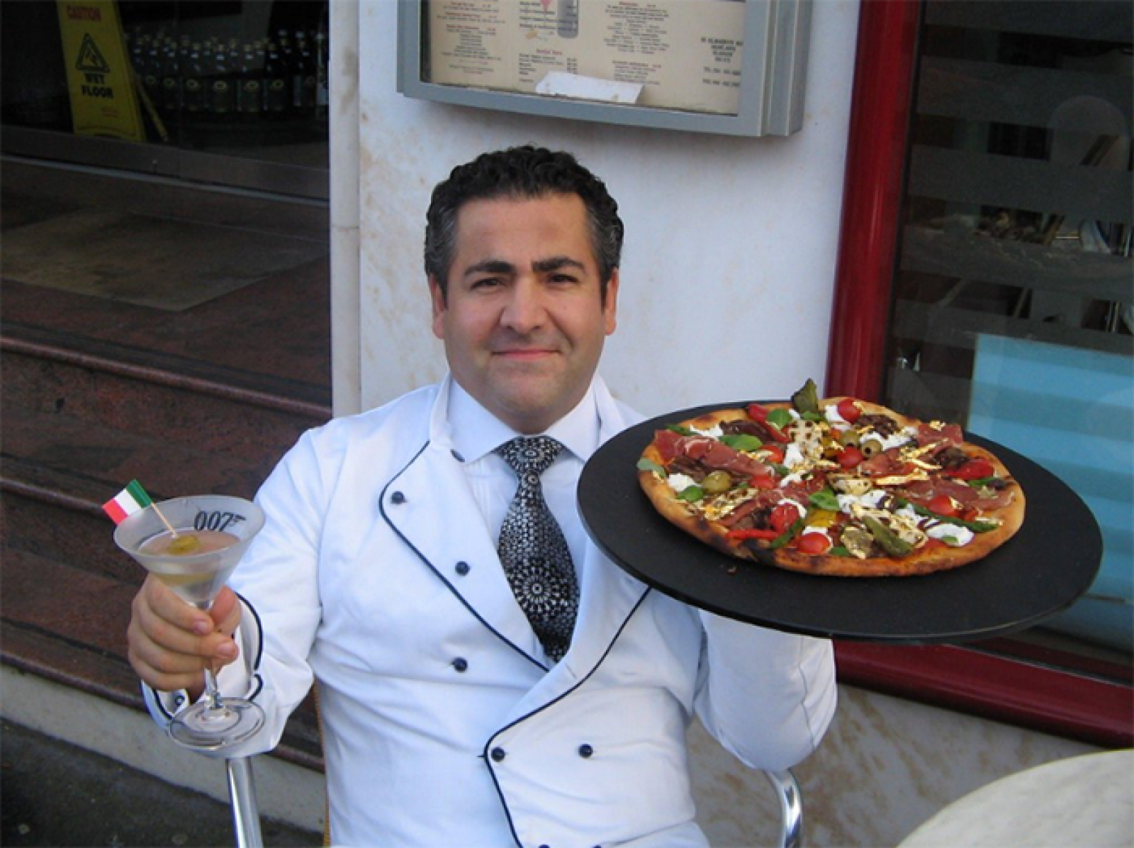 Chef in a white jacket holding a fresh Lebanese-style pizza and a cocktail outside a restaurant
