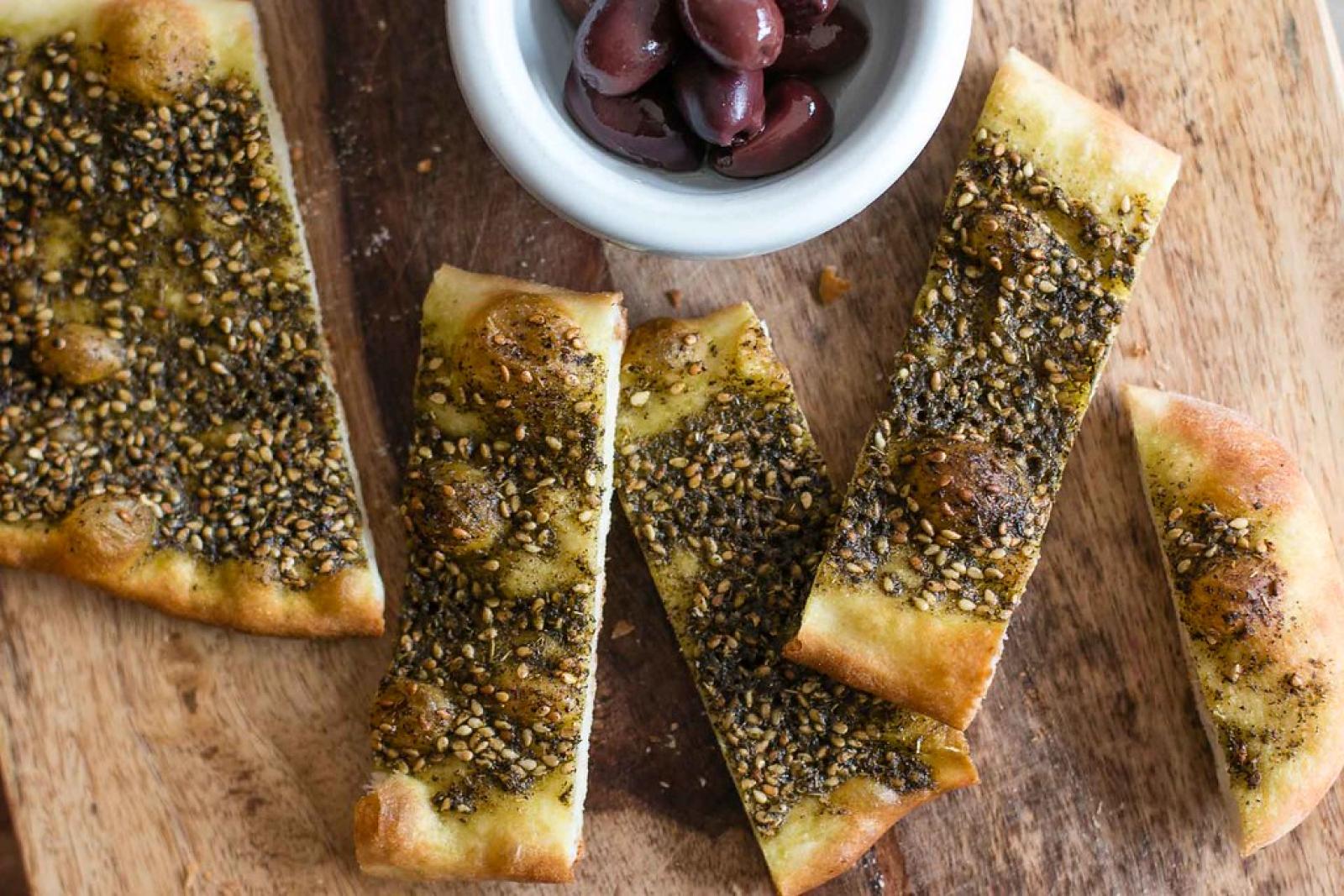 Sliced zaatar manoosh flatbread with a bowl of olives on a wooden board