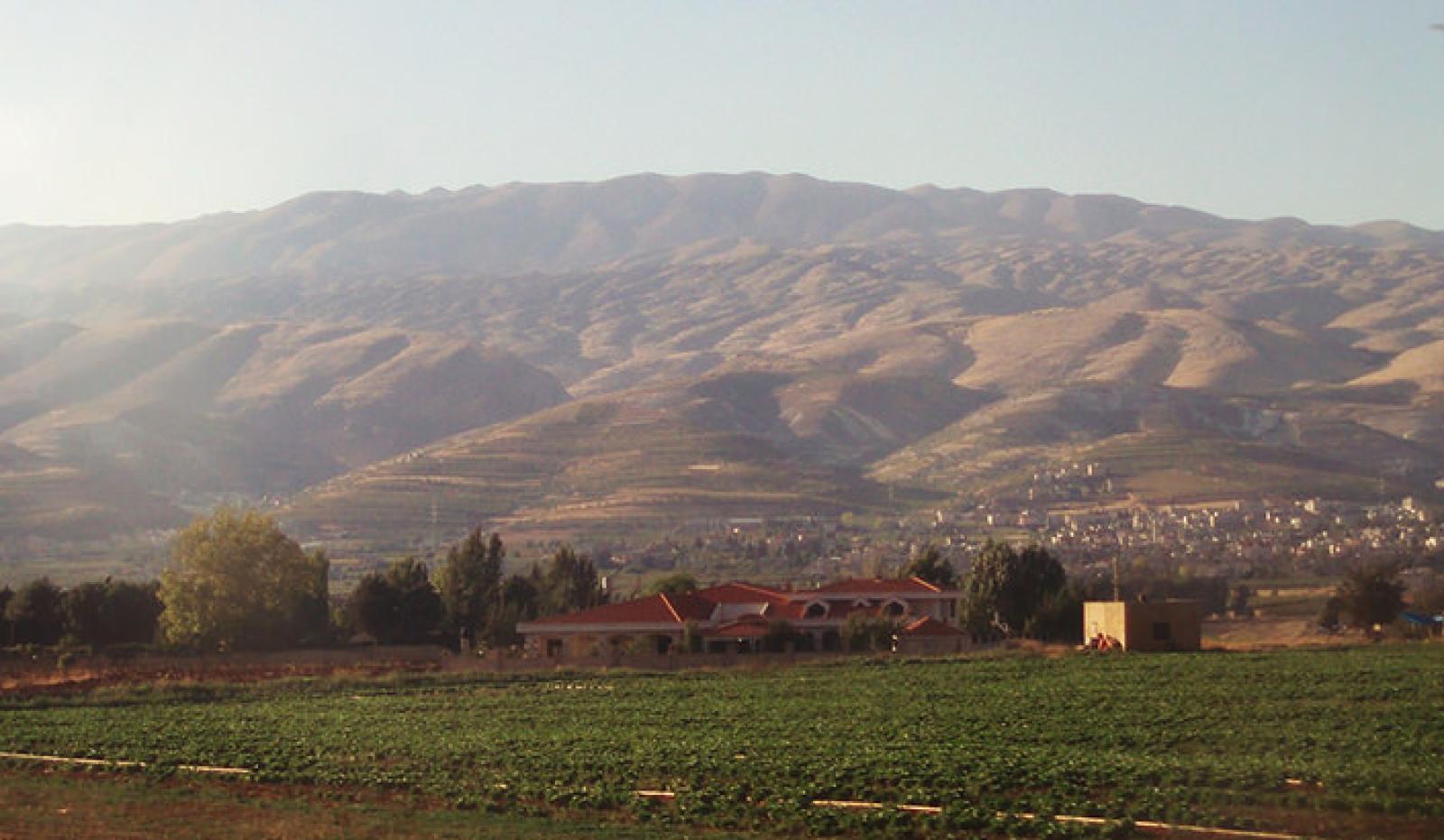 Green farmland and a small village beneath sunlit mountains in rural Lebanon